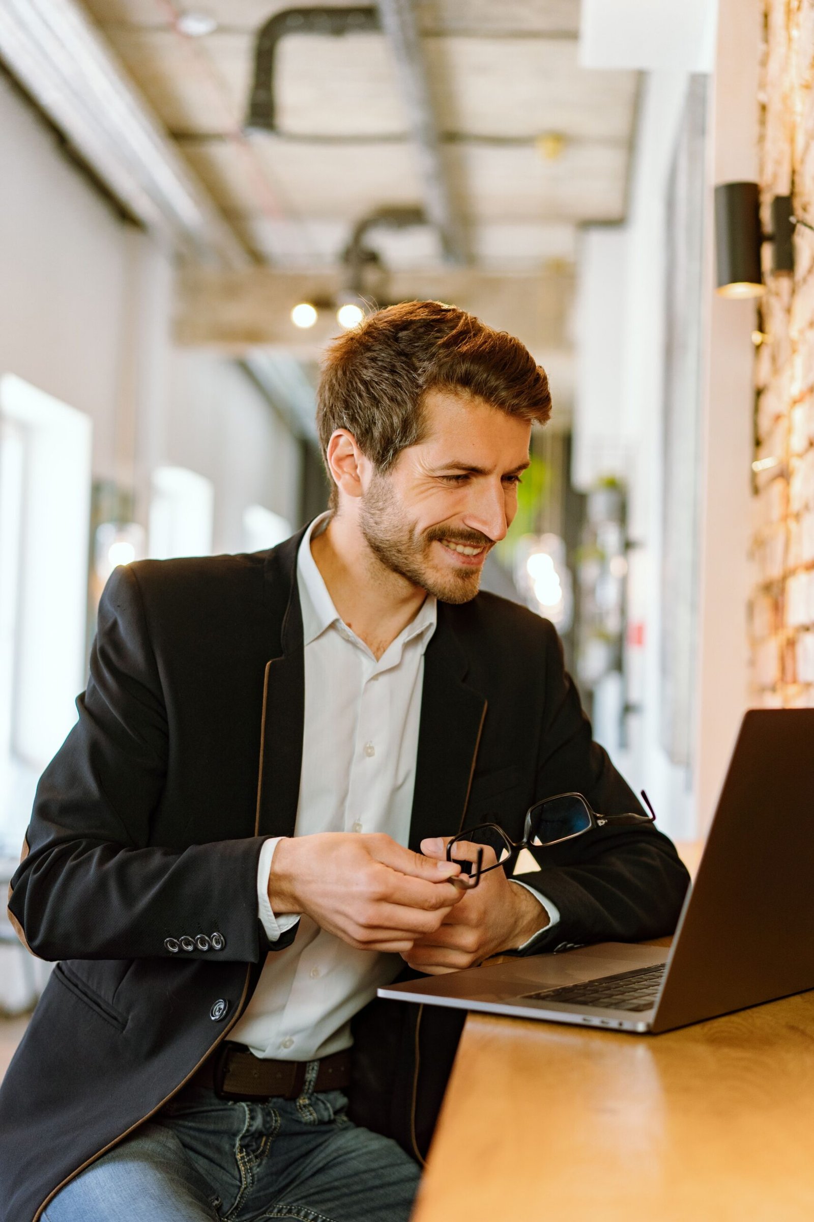 A man in a blazer smiling at his laptop in a bright, tidy workspace; illustrative stock photograph.