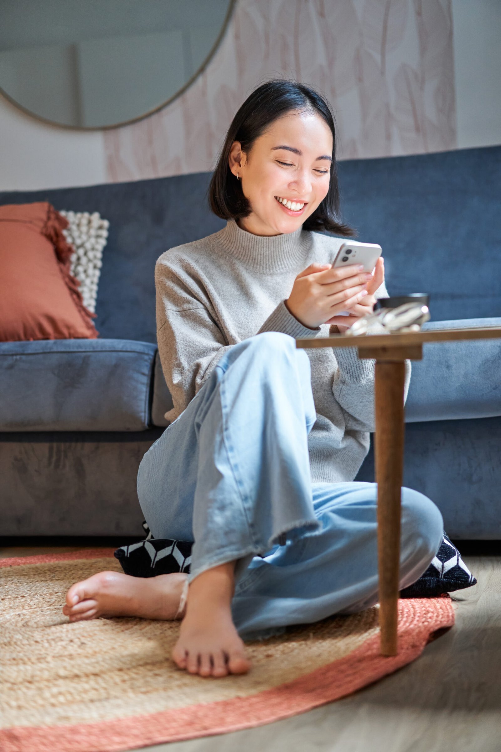 A woman smiling naturally while using her phone in a softly lit living room; illustrative stock photograph.